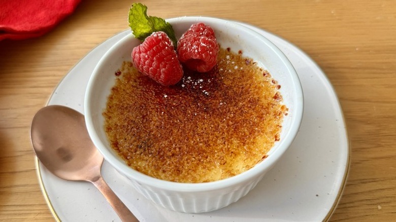 A crème brûlée in a white ramekin topped with raspberries and a mint leaf.