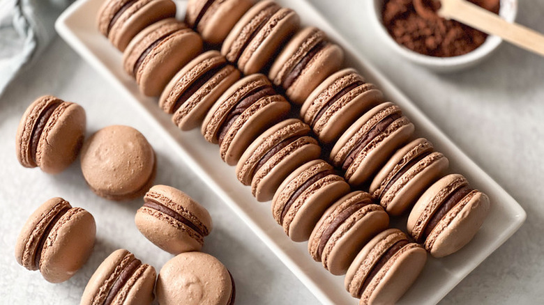 A platter of chocolate macarons next to a bowl of cocoa powder.