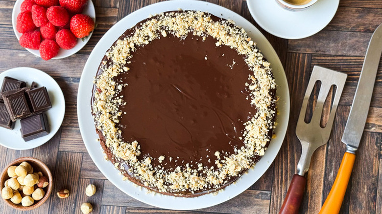 A flourless chocolate cake alongside raspberries, chocolate squares, toasted hazelnuts, and kitchen tools.