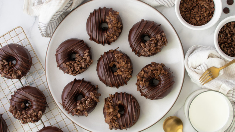 An assortment of chocolate donuts on a plate and wired rack with various tools and ingredients.