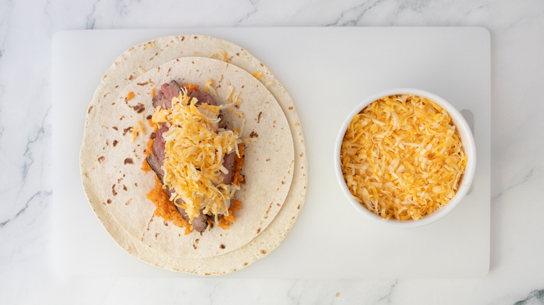 Shredded cheese atop sauce, steak, and rice on top of tortillas