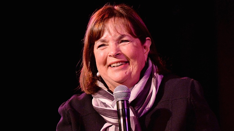 Ina Garten smiling with teeth wearing a grey and white striped scarf and holding a microphone