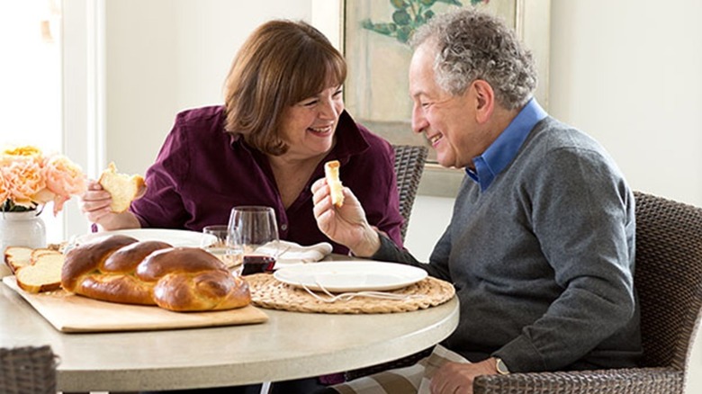 Ina Garten sits at a table with her husband, Jeffrey, as they smile and share a loaf of challah bread