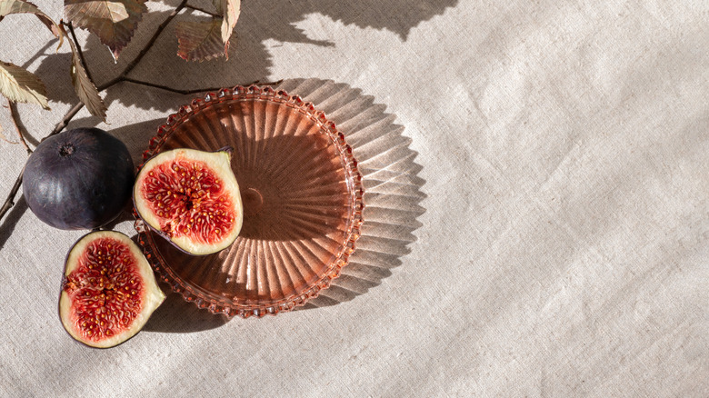 Two fig halves plus a whole fig in a reddish-orange glass dish on a beige tablecloth