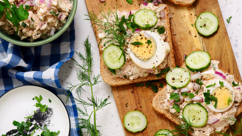 Open-faced tuna salad sandwich with dill, cucumber, and sliced boil egg on cutting board.