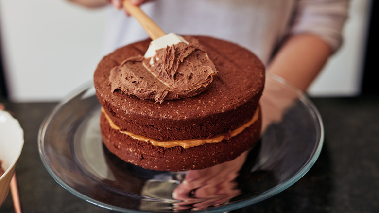 A two-layer chocolate cake is frosted with chocolate frosting with a spatula.