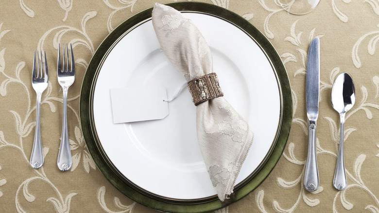 Formal place setting with white plate on green plate, beige napkin in ring on top, and two forks to the left and a knife and spoon to the right