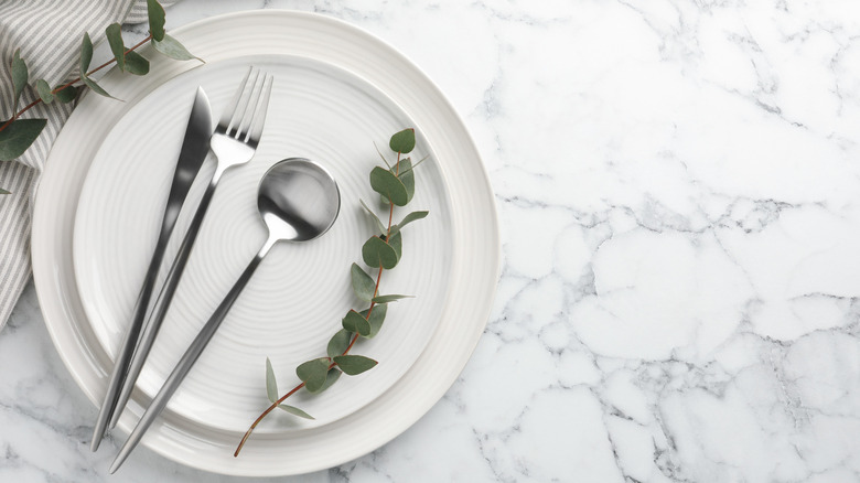 Two white plates stacked together, with a silver fork, knife, and spoon sitting on the top plate next to decorative leaves on a marble surface