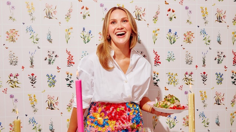Meredith Hayden in a floral apron serving a plate of pasta