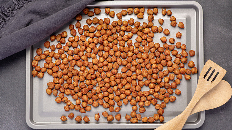 A sheet pan of roasted, spiced chickpeas, partially covered with a tea towel, a wooden spatula and spoon resting on top.