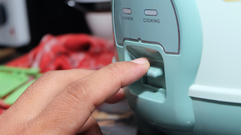 A hand presses the cook button of a teal rice cooker