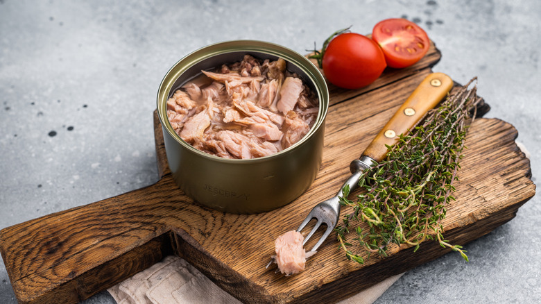 Open can of tuna on a cutting board with herbs and tomatoes