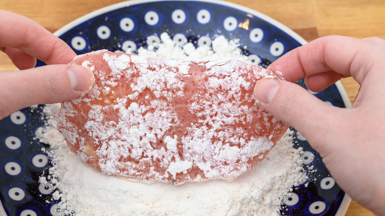Hands dipping a chicken cutlet into flour.