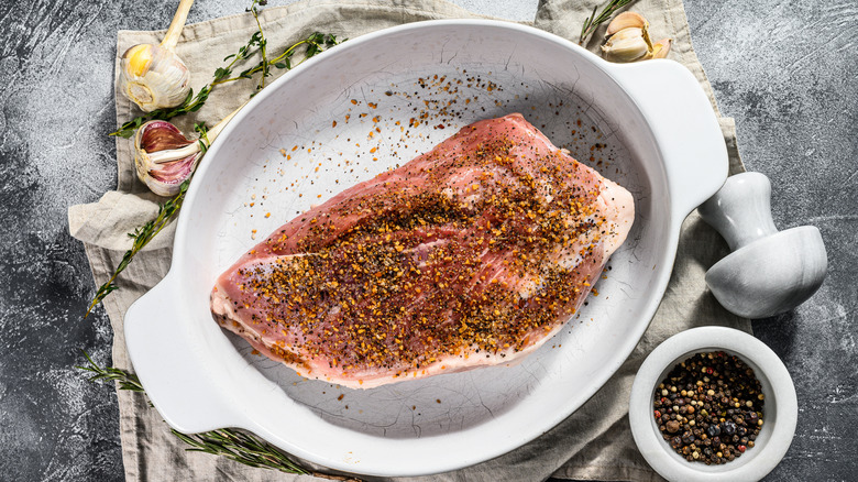 Pork shoulder in white roasting pan covered in spices and surrounded by garlic and herbs