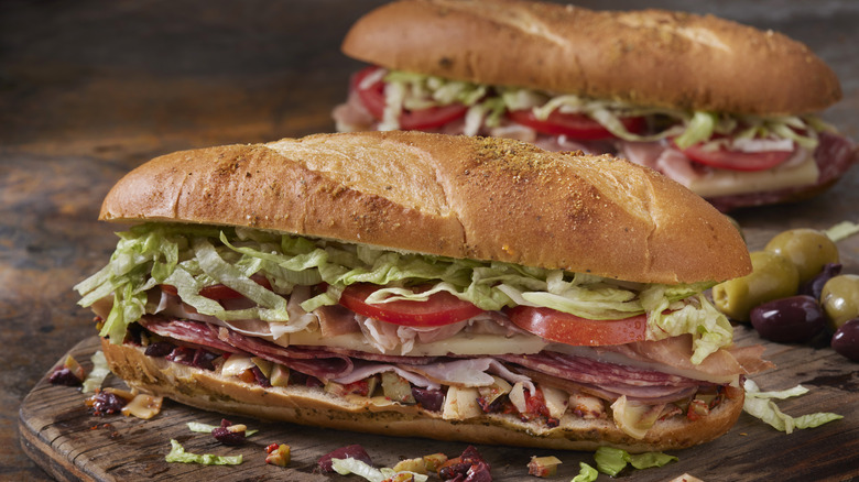 Itlalian sub with vegetables, cold cuts, tomatoes, and lettuce on a wooden cutting board.