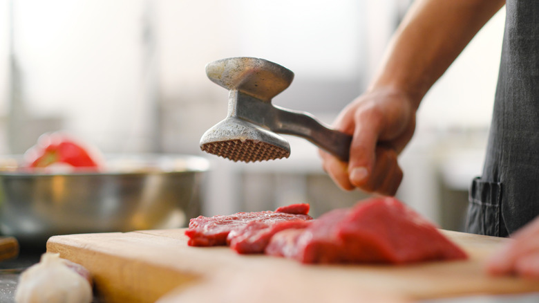 A cook tenderizing a piece of meat on a cutting board with a meat mallet