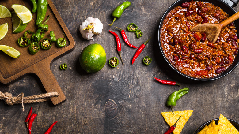 Chili is in a pan with a wooden spoon; chilis, garlic, and limes are also styled in the frame