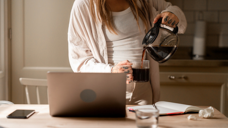 A woman pours a cup of coffee in a home setting, with a laptop and a notebook on the table.