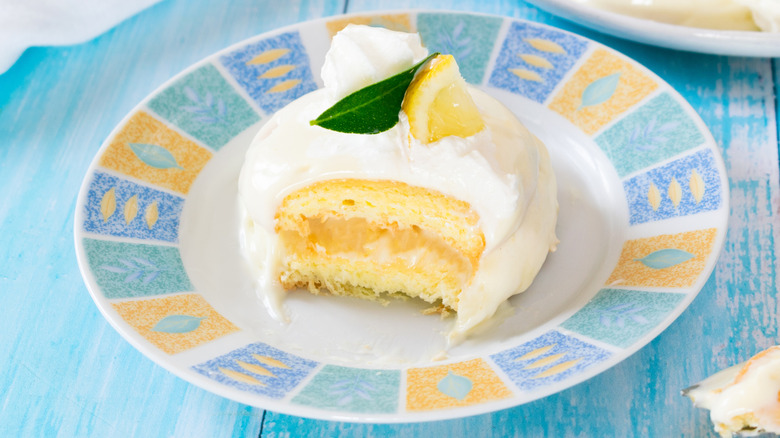 Delizia al limone or lemon delight garnished with a lemon wedge on a colorful plate. A forkful has been removed, revealing a cross-section with layers of cream, cake, and filling.