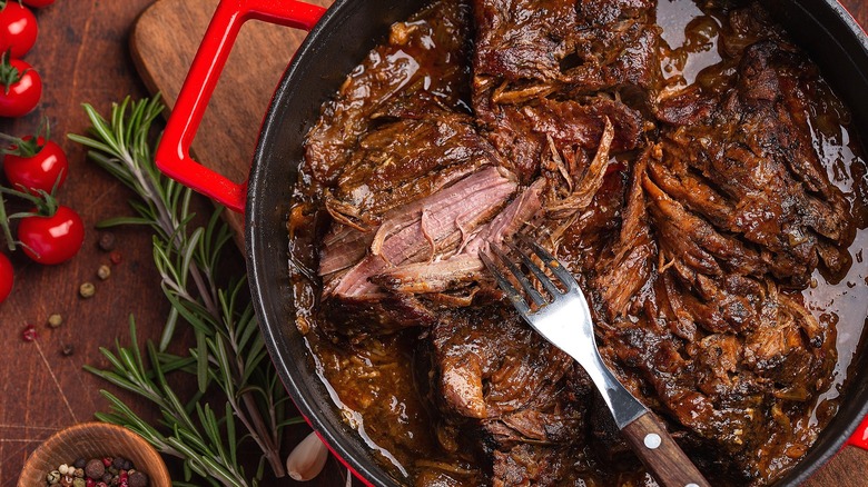 Slow-cooked pot roast in a red-enamel pot surrounded next to a bunch of cherry tomatoes and fresh rosemary.