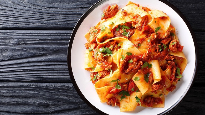 Stracotto mixed with pappardelle pasta and green herbs on a white plate with black trim.