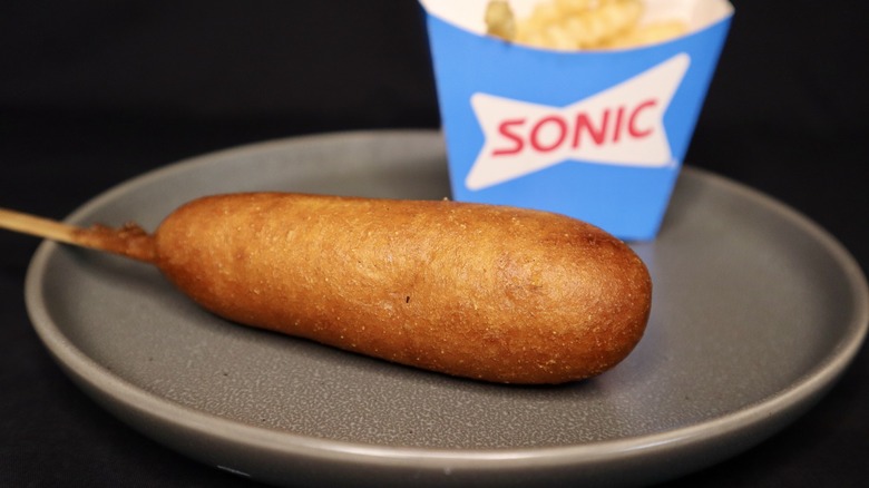 a single corn dog on plate with carton of Sonic fries in background