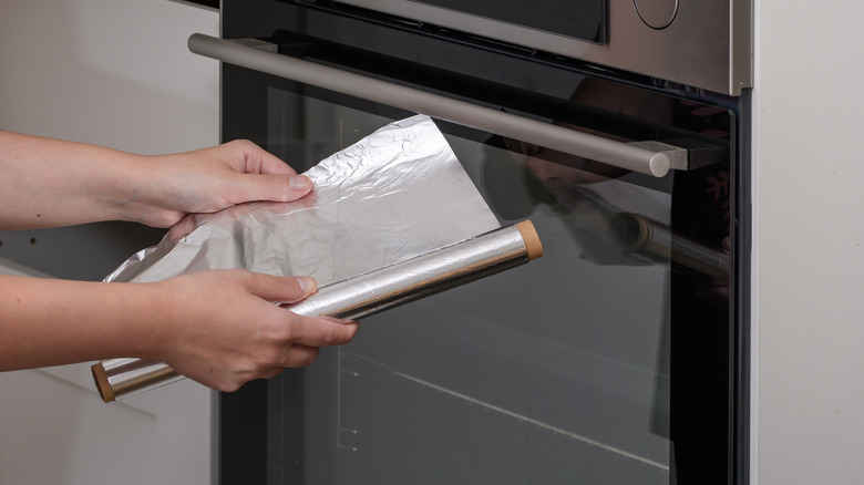 A person unrolling a sheet of aluminum foil in front of a closed oven.