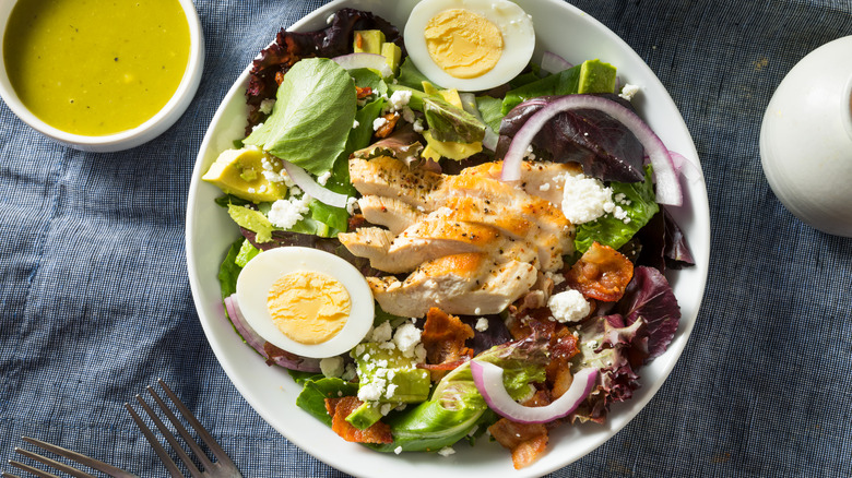 A large salad with mixed greens, onions, avocado, goat cheese, two egg halves, and sliced chicken breast with a ramekin of green goddess on the side.