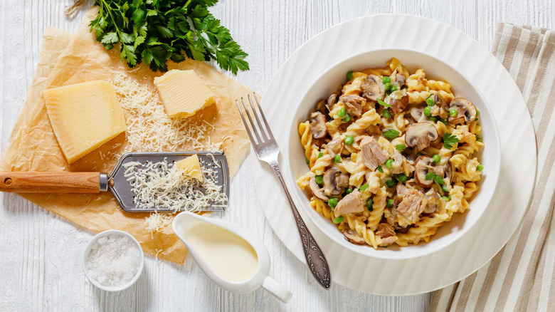Pasta with tuna chunks, mushrooms, and peas in white bowl on a table beside Parmesan cheese
