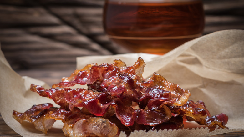 Freshly cooked bacon rests on baking paper in front of glass of whiskey in background.