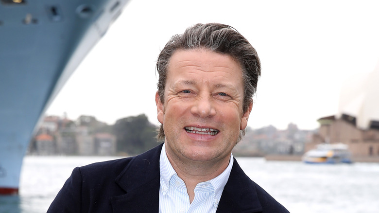 Jamie Oliver smiling in Sydney, Australia, standing on a dock in front of a large ship