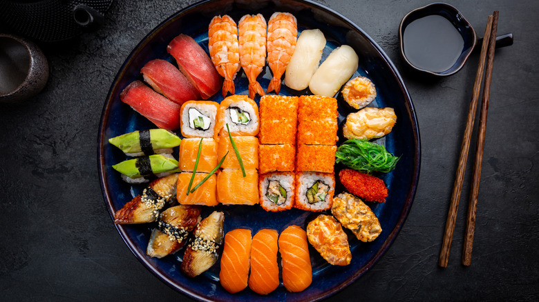 Plate of sushi rolls and sashimi on a brown and blue plate next to chopsticks and cup of soy sauce