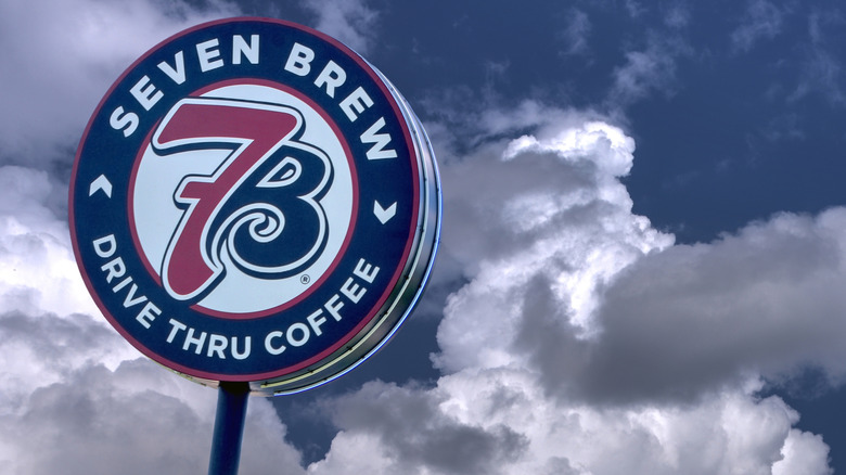 A circular sign with the 7 Brew logo pictured against a cloudy sky. The sign reads: "Seven Brew drive thru coffee"
