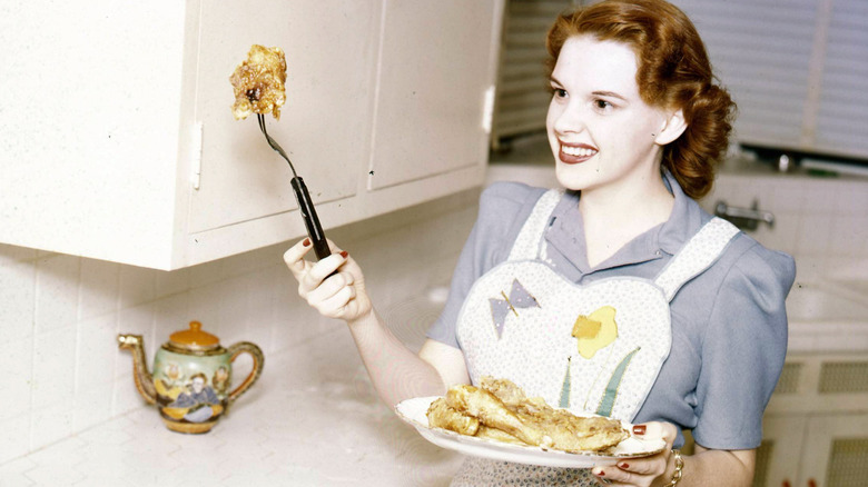 Judy Garland standing in a kitchen with a plate of chicken in one hand and a long-pronged fork with a piece of chicken on the end in the other hand.