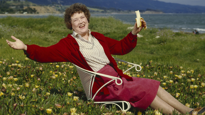 Julia child sitting in a white metal chair in a field of flowers with a banana in her hand