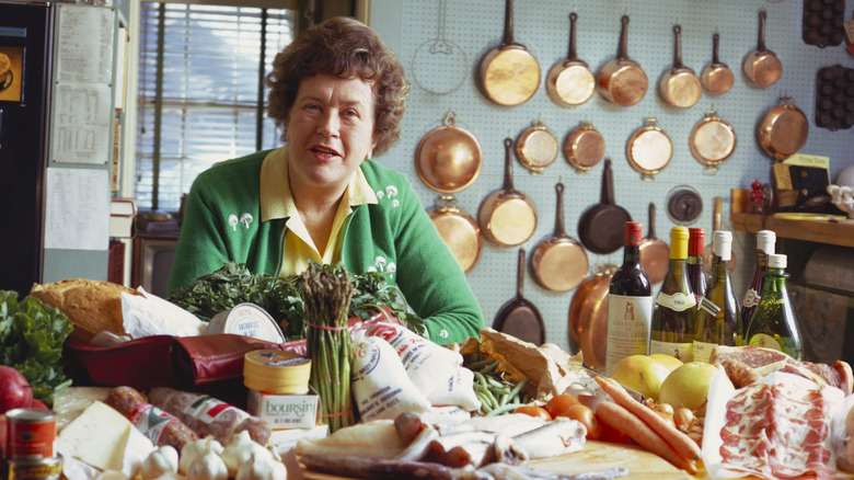 Julia Child in a kitchen with groceries on the counter in front of a wall of copper pans