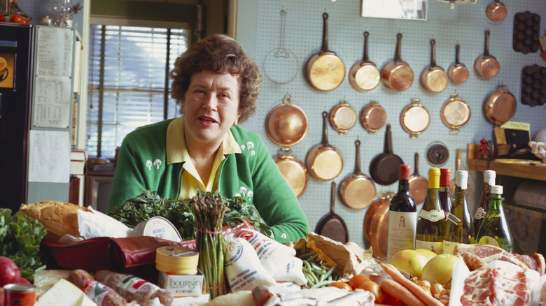 Julia Chiild appears at a table abundant with produce and wine bottles.