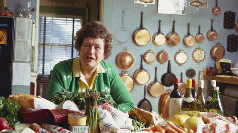Celebrity chef Julia Child sits at a kitchen table with mounted copper pots on the wall behind her.