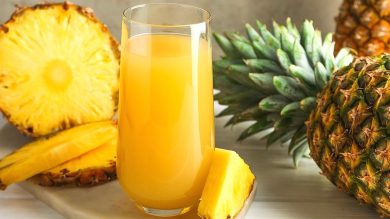 Clear glass of pineapple juice surrounded by whole and cut pineapples