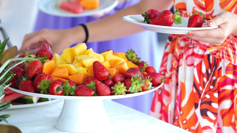 Woman taking fruit off a white elevated platter filled with whole strawberries and cut cantaloupe and pineapple