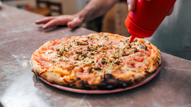 Person drizzling ketchup from a squeeze bottle over a cooked pizza