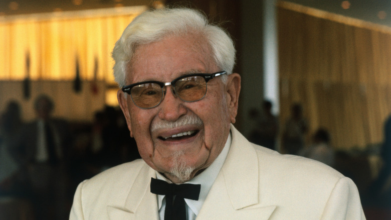 The real Colonel Harland Sanders smiling while wearing his iconic white suit and tie.