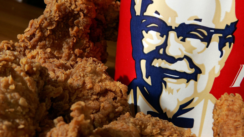 KFC fried chicken pieces in front of a bucket with Colonel Sanders' face on it.