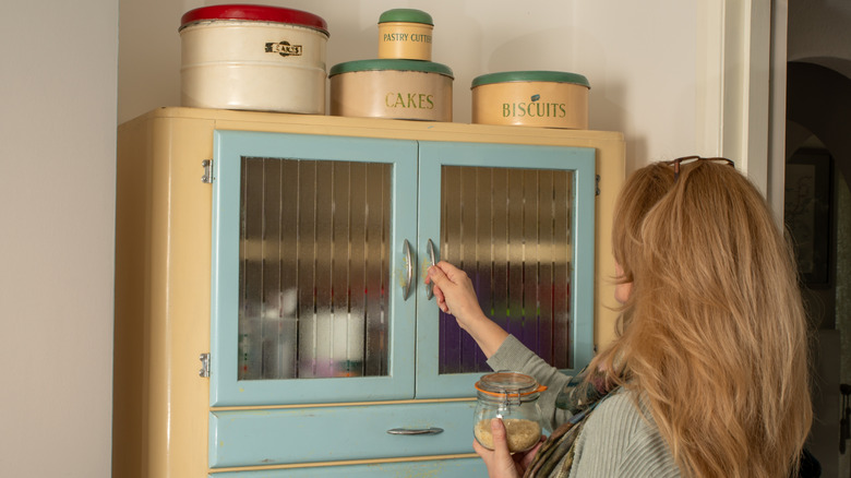 Woman putting away a jar of dry goods into a freestanding blue and yellow old school larder