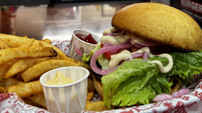 A close-up of a burger piled with toppings, served alongside fries, ketchup, and mayo