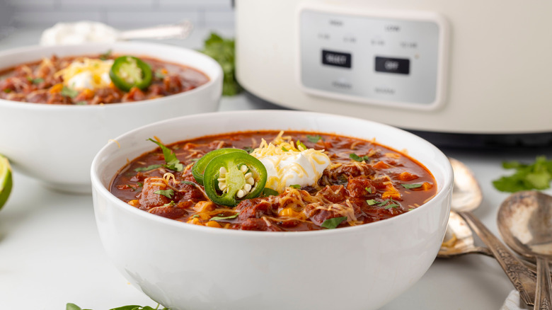 two white bowls of chili with jalapenos and sour cream on top, beside white slow cooker