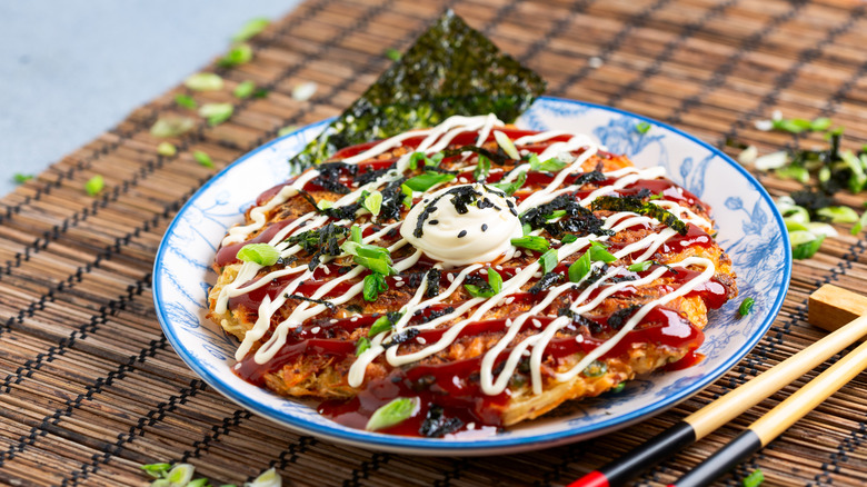 an okonomiyaki garnished with various sauces and seasoning on a blue plate next to chopsticks