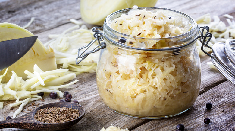 an opened jar of sauerkraut with various dried spices and sliced cabbage beside it