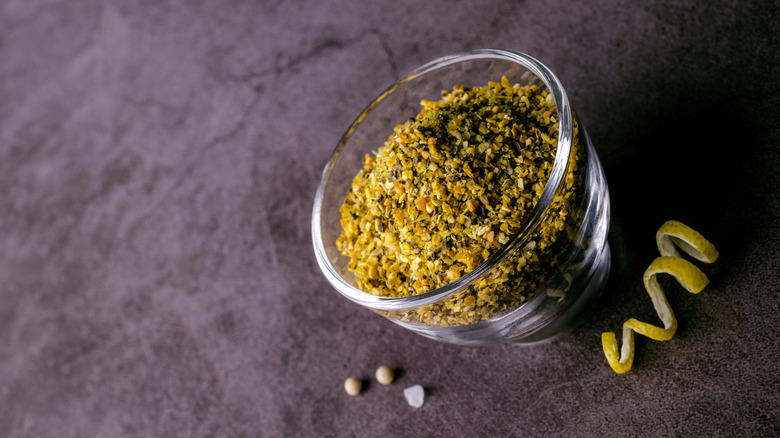 A clear cup of lemon pepper seasoning on a tabletop with a spiraled lemon peel, peppercorns, and a grain of salt