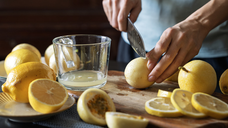Person slicing and juicing fresh lemons into clear cup on wood cutting board.